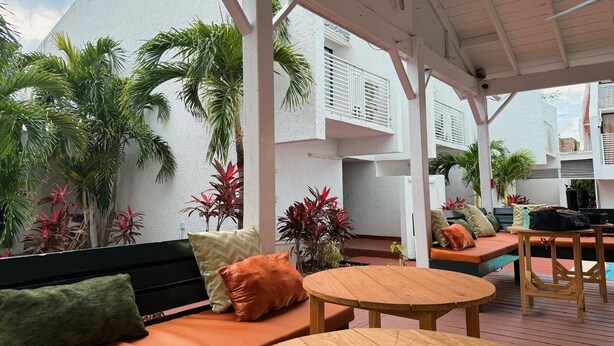 Un espacio de estar al aire libre cubierto con muebles de madera, un edificio blanco al fondo y plantas tropicales.