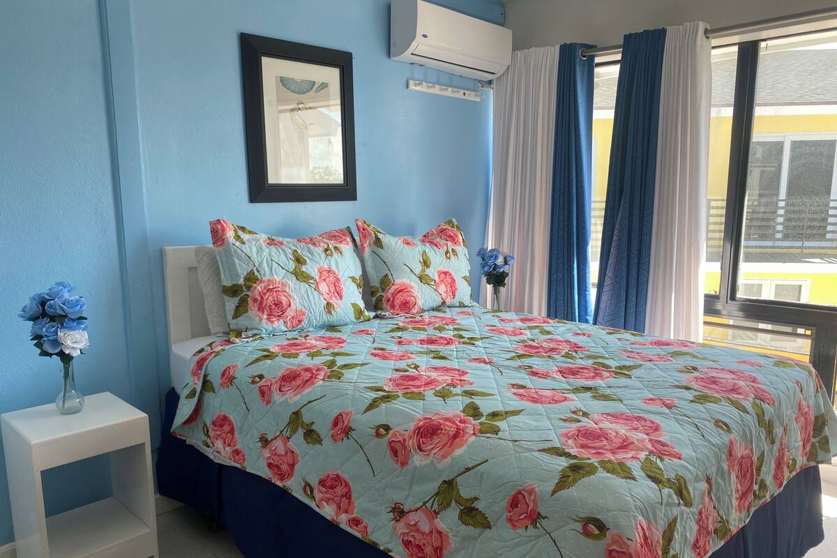Un dormitorio con colcha estampada, una mesita de noche blanca con un florero y un cuadro en la pared.