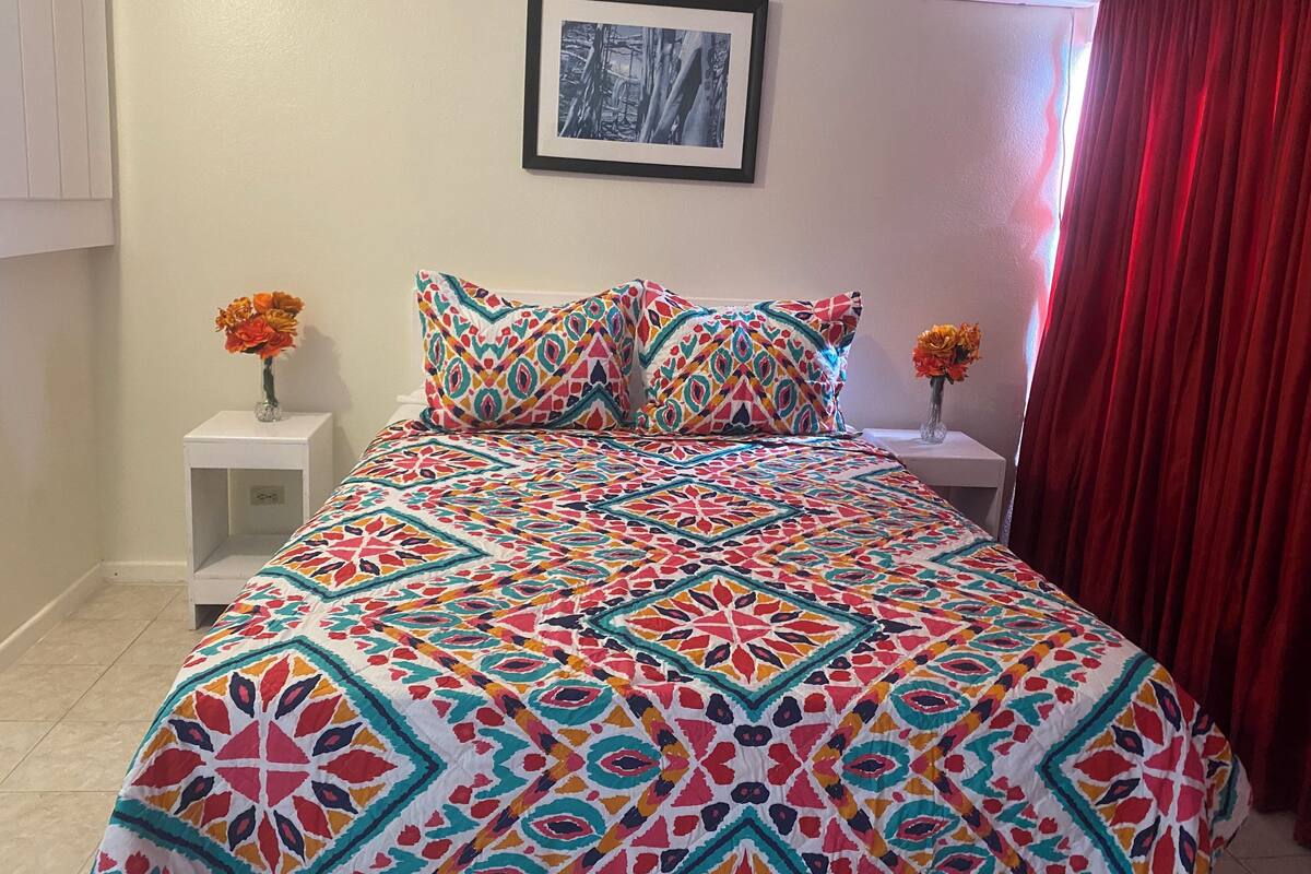 Una cama con un edredón estampado y colorido, dos mesitas de noche blancas con jarrones de flores, un cuadro en la pared y una cortina roja.