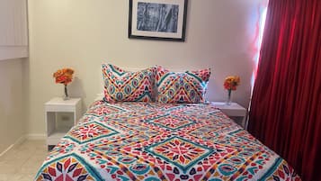 A bed with a colorful patterned bedspread, two white bedside tables with flower vases, a framed picture on the wall, and a red curtain.
