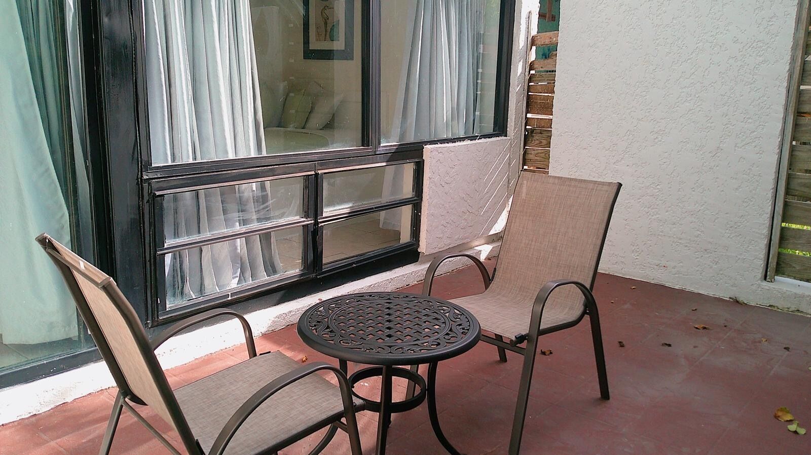 Eine Terrasse mit Tisch und zwei Stühlen, ein Fenster mit Vorhängen und eine Ziegelwand.