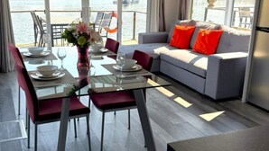 Un salón moderno con una mesa de comedor de cristal, sillas rojas, un sofá gris con cojines naranjas y vistas a un puerto deportivo a través de amplias ventanas.