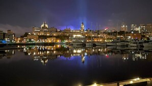 Un paisaje urbano nocturno con un puerto deportivo y barcos, un flotador en una barandilla y luces colgantes.