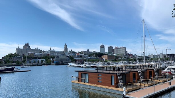 Un puerto deportivo con barcos amarrados, un edificio moderno con revestimiento de madera y un paisaje urbano al fondo.