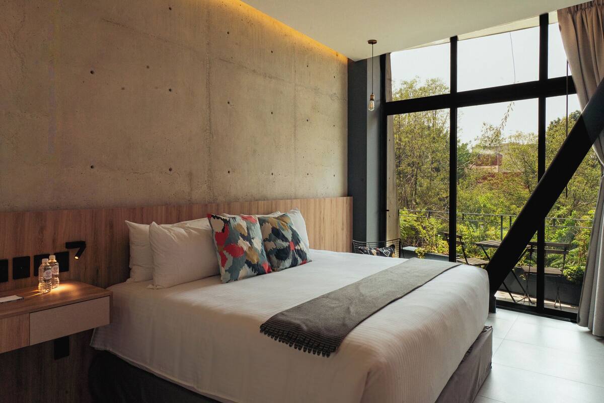 A modern bedroom with a large bed, wooden headboard, and a view of greenery through large windows.