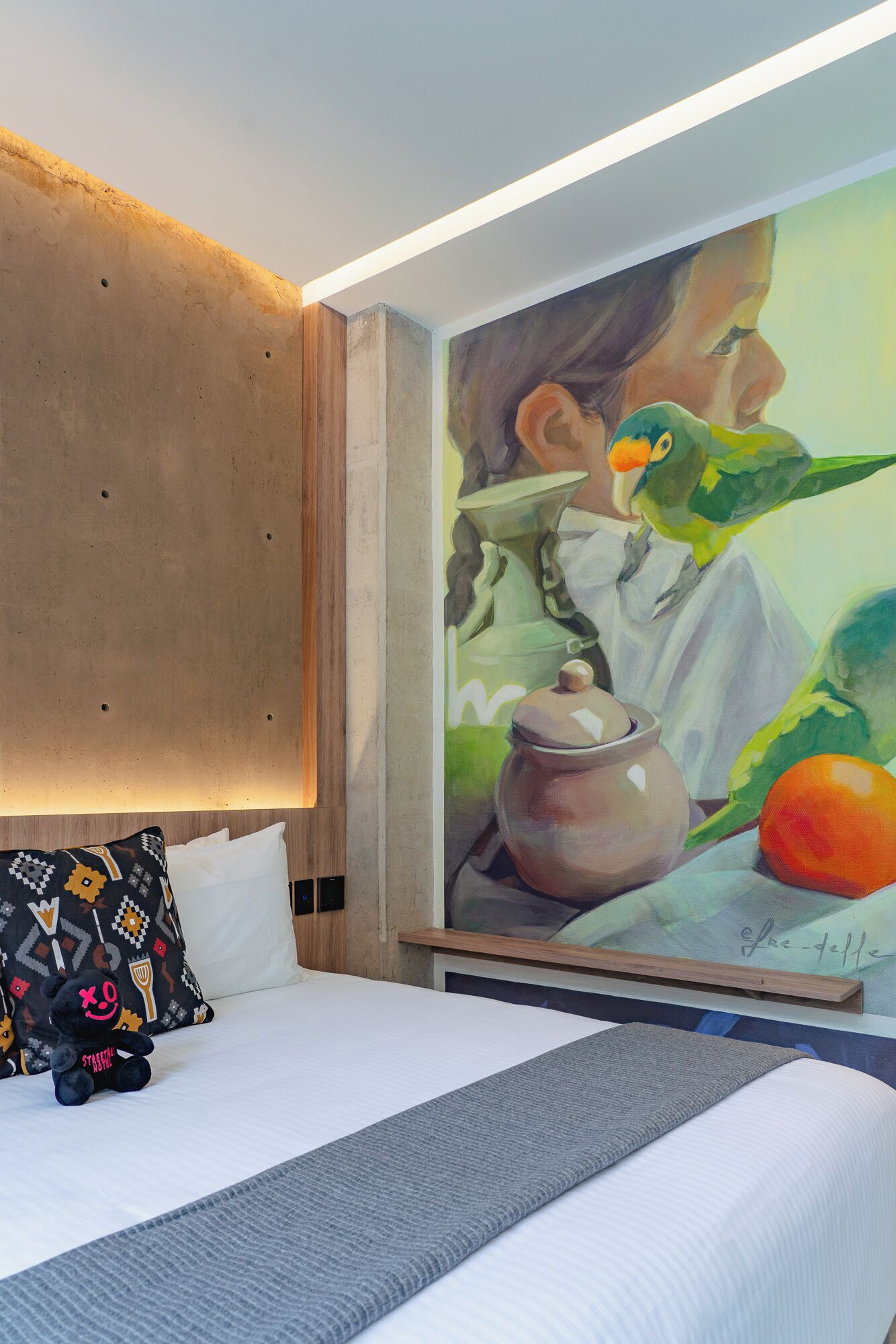 Quarto de hotel com cama, pintura de uma mulher e um pássaro, e travesseiro em formato de urso.