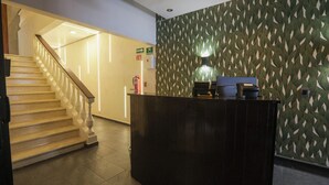 Ein dunkler Empfangstresen mit einer gemusterten Wand dahinter. Eine Treppe mit Holzbalkon ist links zu sehen.