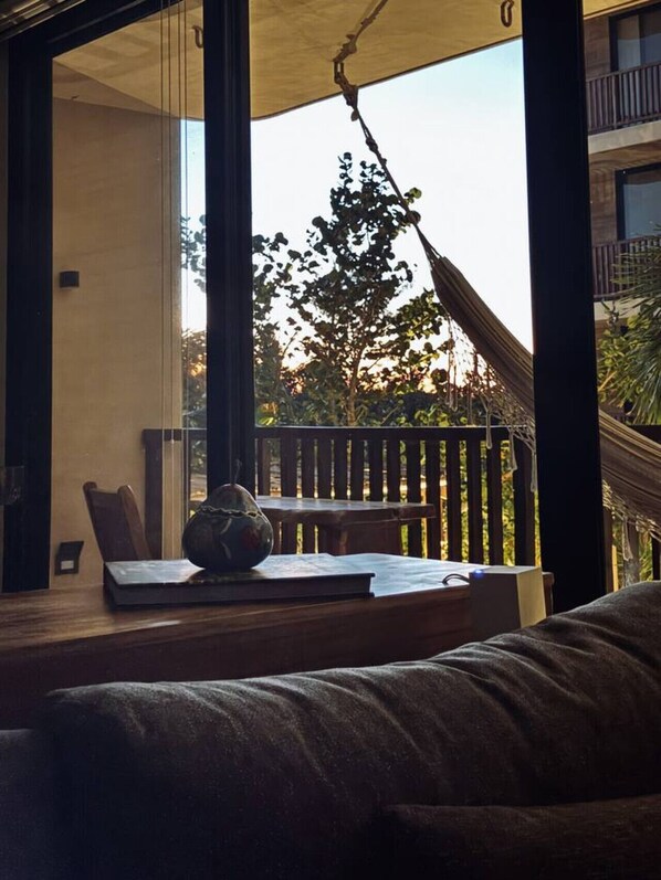 Una habitación con un ventanal grande, una hamaca, una mesa de madera y un rincón de estar con cojines.