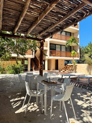 Un área de estar al aire libre con sillas y mesas de plástico blanco, un pérgola de madera sobre ella y un edificio con balcones al fondo.