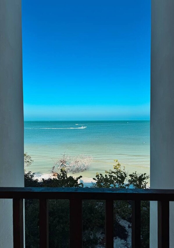 A view from a balcony overlooking a beach with clear blue water and a boat in the distance.