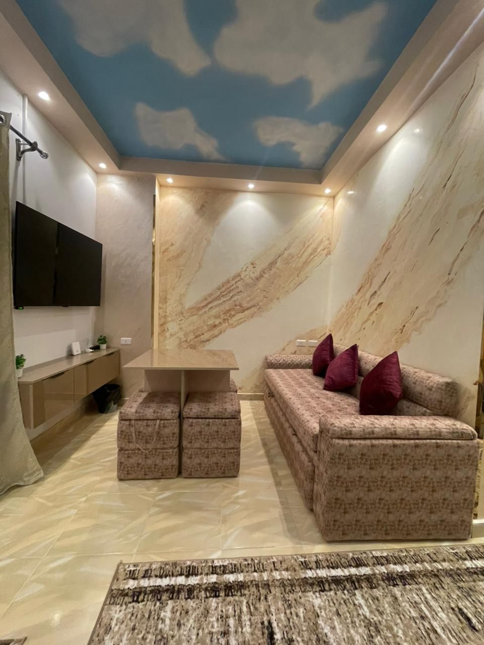 A modern living room with a marble-patterned wall, a flat-screen TV mounted on the wall, a sofa with cushions, and a coffee table.