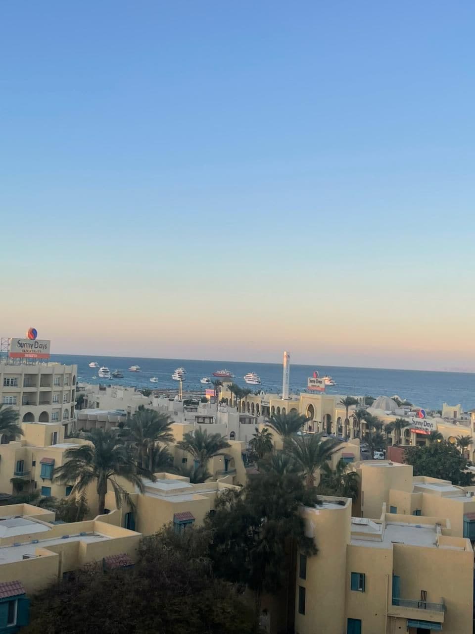 Un pueblo costero con edificios que presentan ventanas arqueadas y balcones, una torre destacada y vista al mar con barcos.