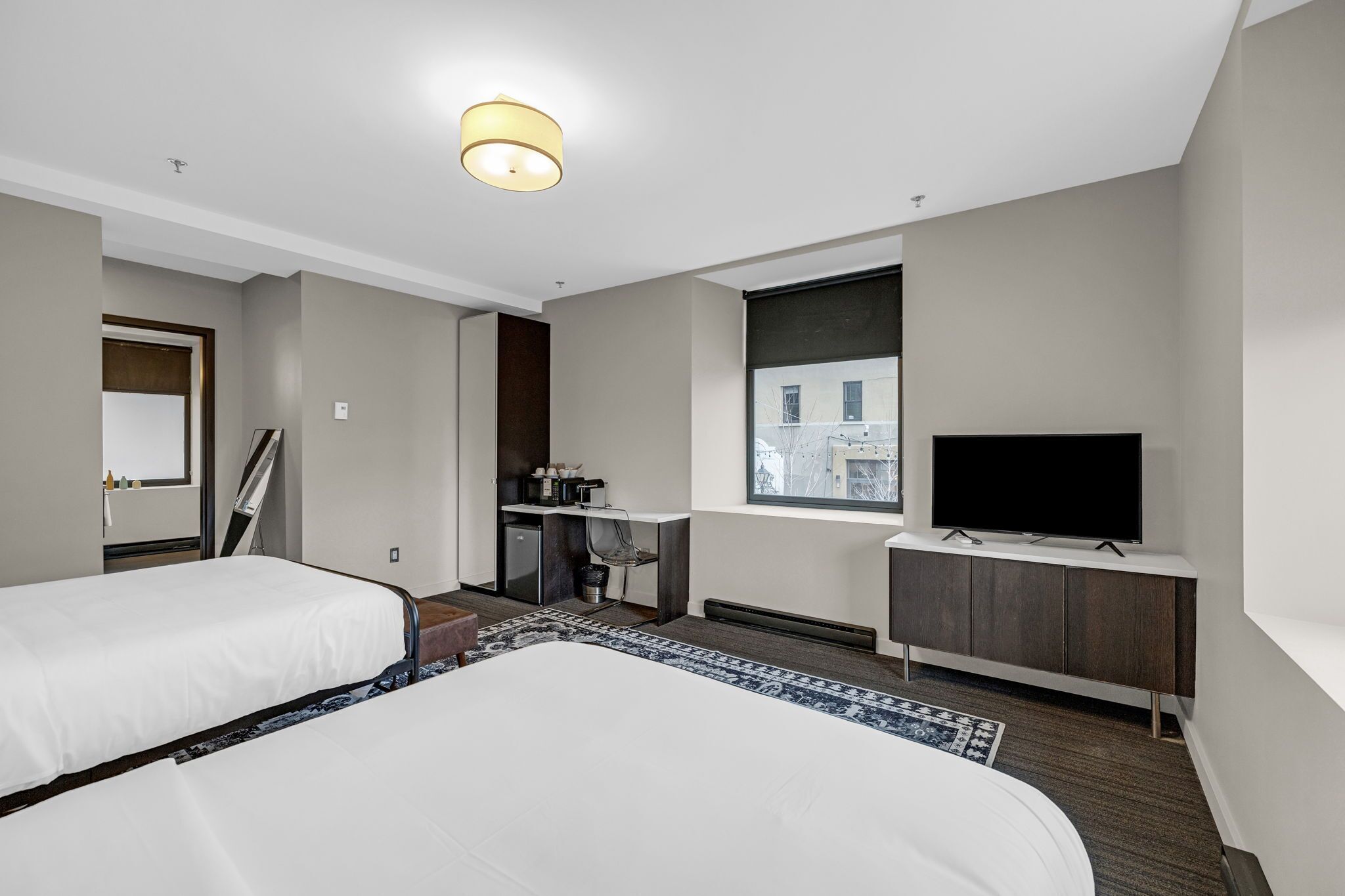 Habitación de hotel con dos camas, un escritorio con silla, televisión y ventana con vista a edificios.