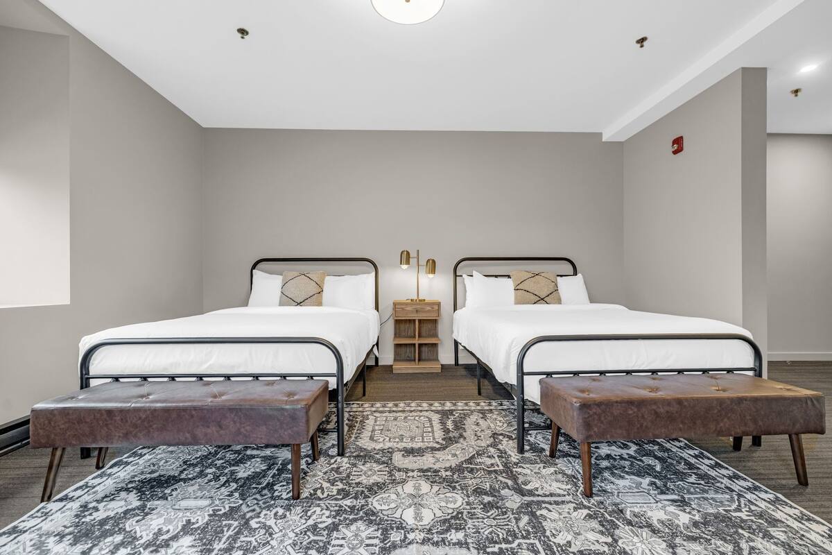 A modern hotel room with two beds, a central nightstand, and a patterned rug.