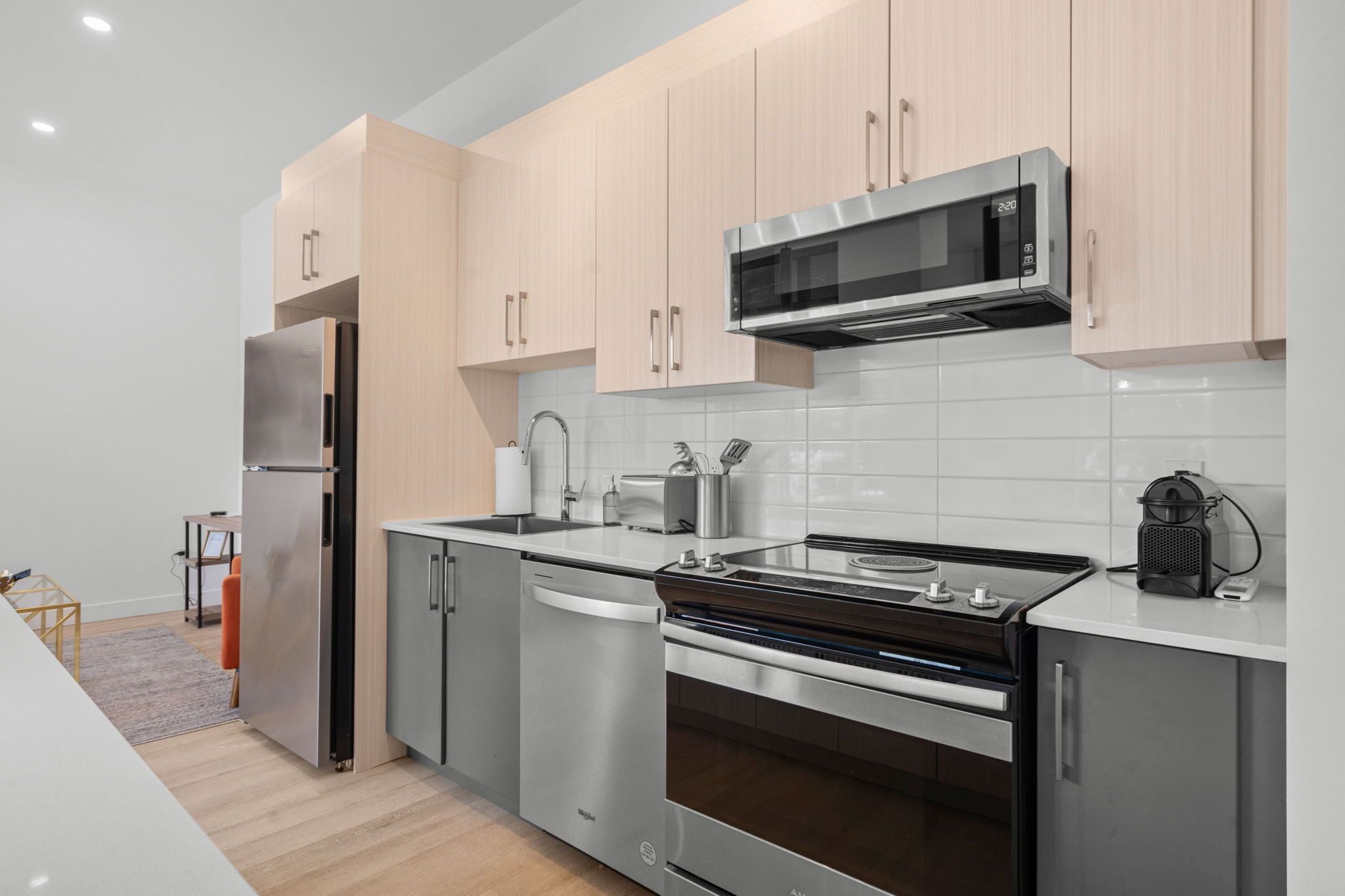 A modern kitchen with stainless steel appliances, light wood cabinets, and a white tiled backsplash.