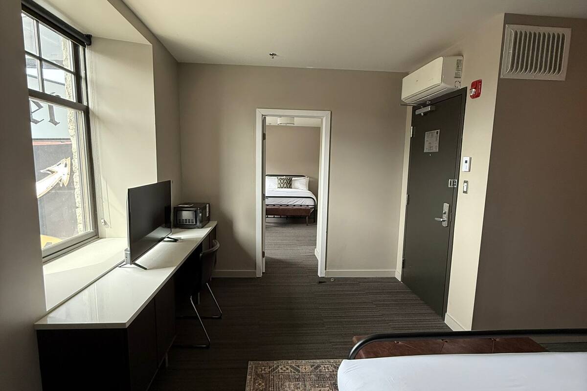 A hotel room with a bed, desk, and window. There is a door leading to another room and a vent on the wall.
