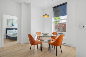 Una habitación moderna con una mesa de comedor de vidrio, sillas anaranjadas, una cama con cabecera azul y un ventanal con vistas a edificios.