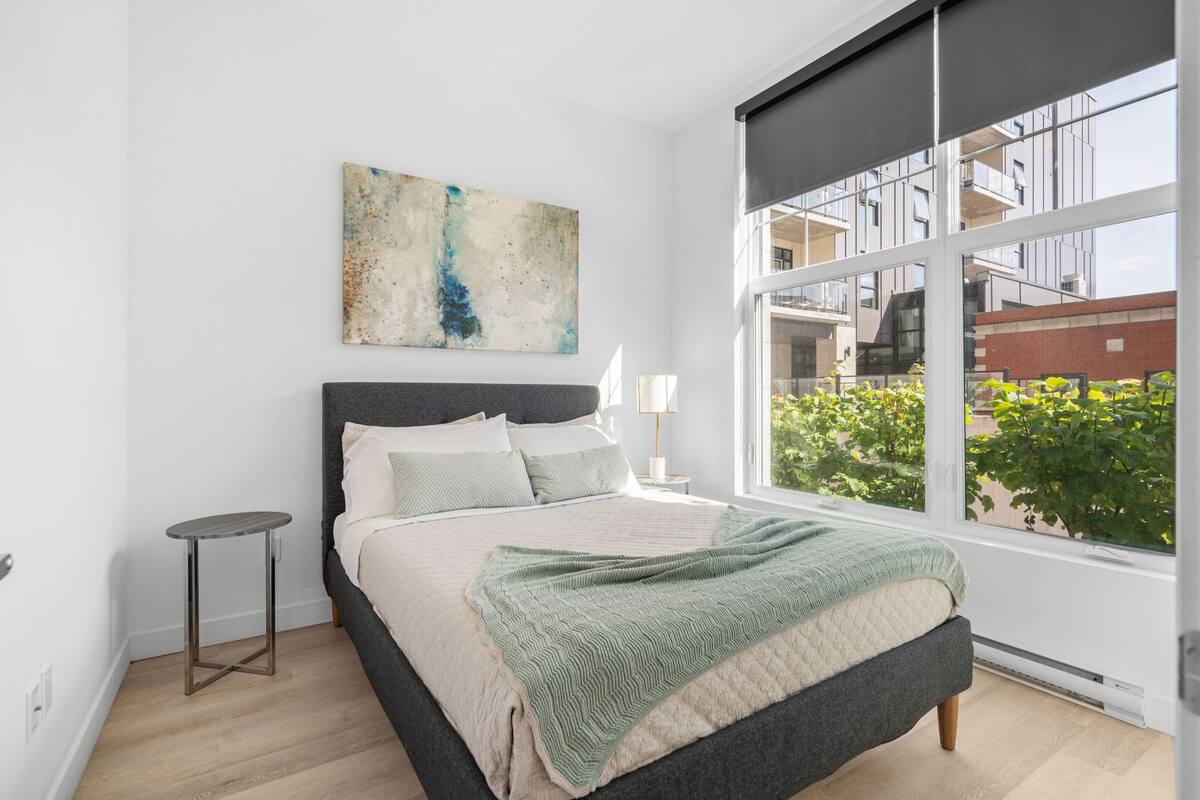 A modern bedroom with a large window, a bed with a green blanket, and a small side table.