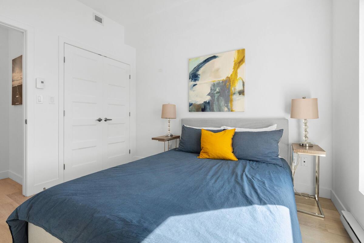 A bedroom with a large bed, two bedside tables, a painting on the wall, and a white door.