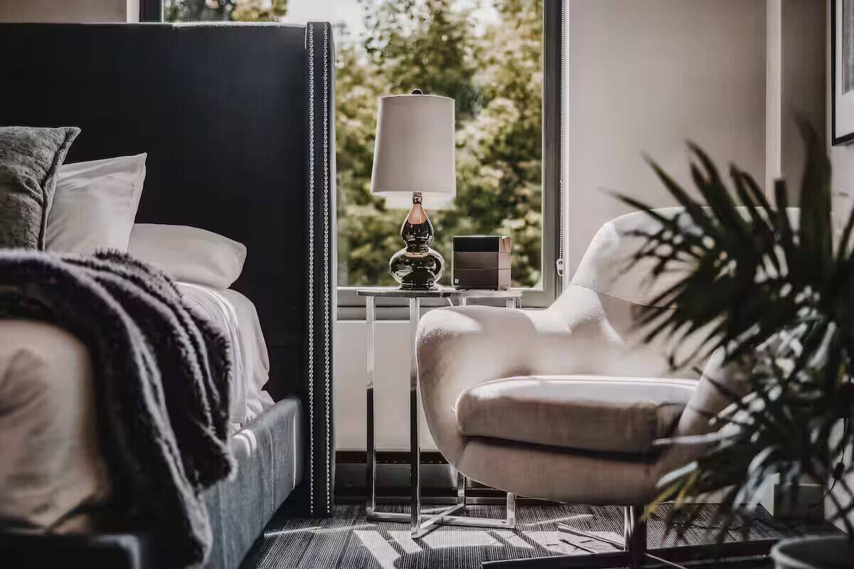 A modern bedroom with a large bed, a bedside table with a lamp, a chair, and a view of greenery outside.