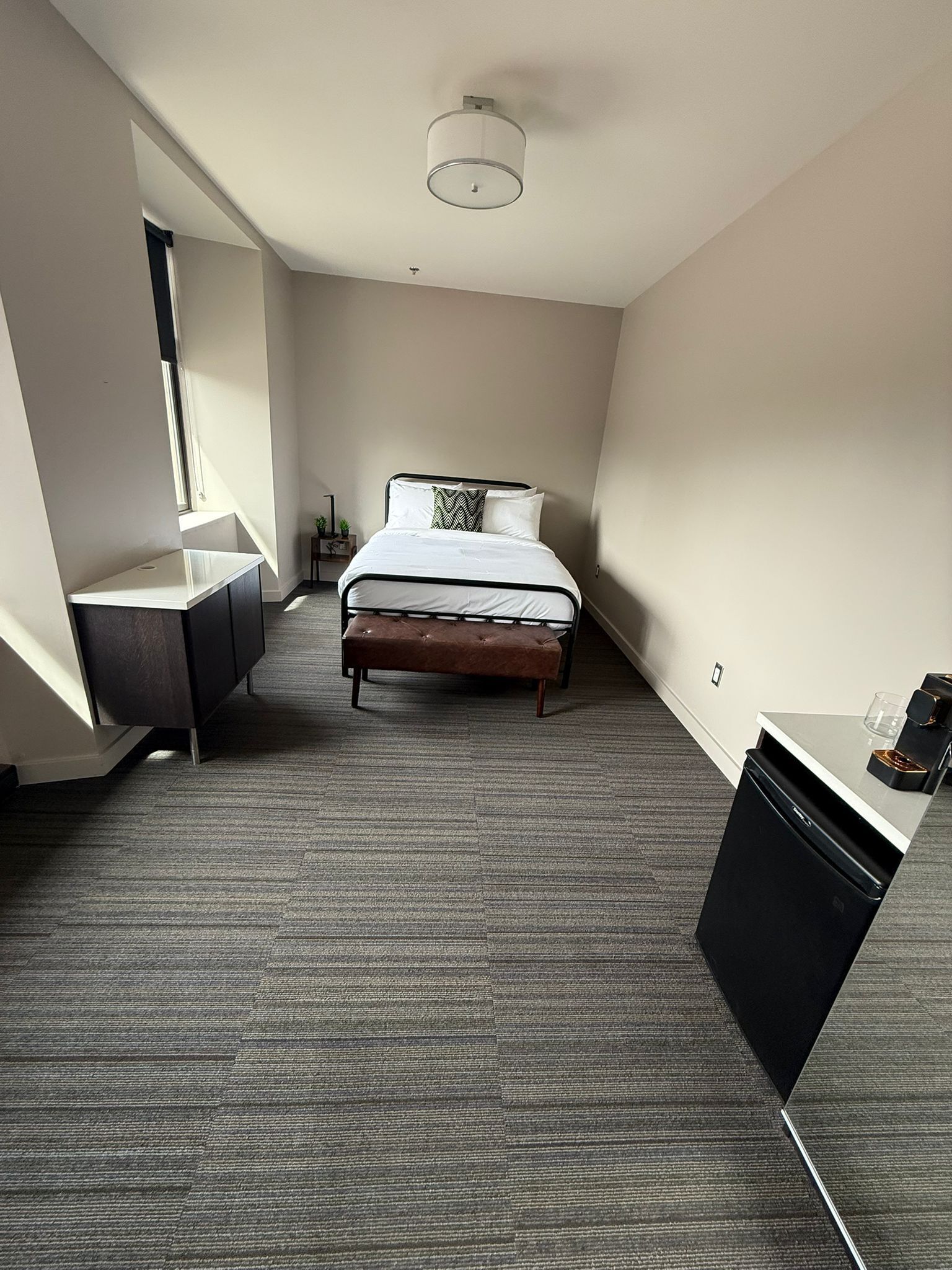 Una habitación de hotel moderna con cama, un escritorio pequeño, una silla y una kitchenette compacta.