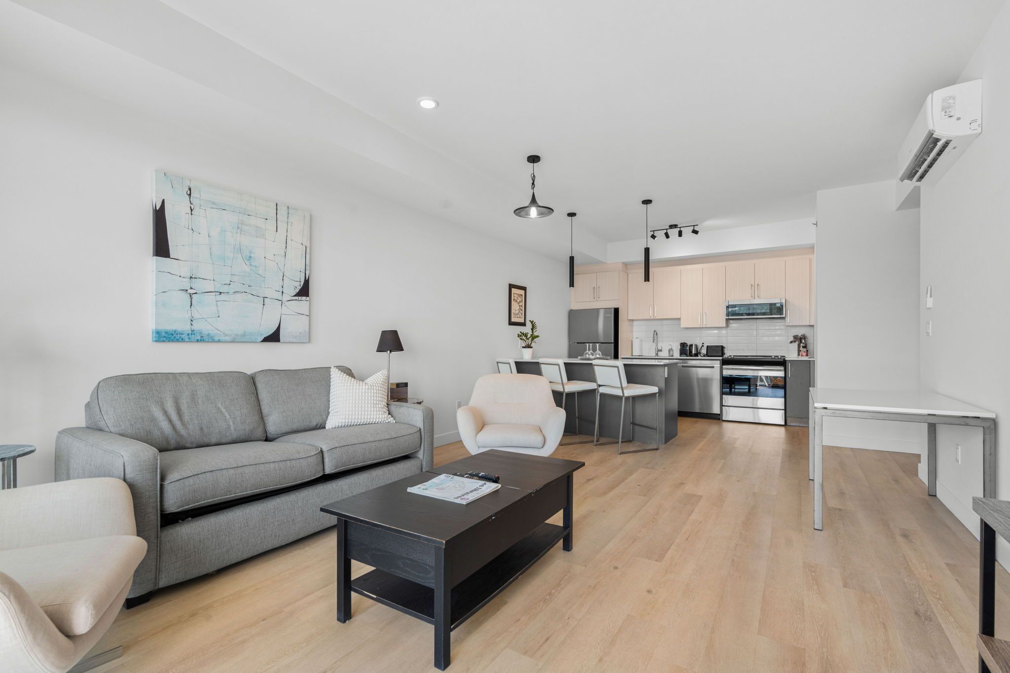 Un salon moderne avec un canapé gris, une table basse noire et un fauteuil blanc. On aperçoit une cuisine à l’arrière-plan, avec un coin repas.