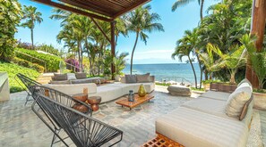 A covered outdoor seating area with a sofa, chairs, and a coffee table, overlooking a beach with palm trees and the ocean. - Cruz de Huanacaxtle Arena Blanca 408 (La Cruz de Huanacaxtle)