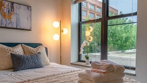 Een net opgemaakt bed met een gebreid plaid, een bedlampje en een raam met uitzicht op een gebouw en bomen.