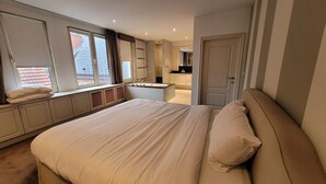 A bedroom with a large bed, a built-in window seat, a bathroom with a shower, and a kitchen area visible through an open door. - Spacious 3BR apartment with rooftop terrace (Antwerpen)