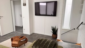 Ein modernes Wohnzimmer mit einem grünen Sofa, einem Holztisch, einem an der Wand montierten Fernseher und einer Zimmerpflanze.