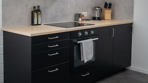 Eine moderne Küche mit einem schwarzen Herd, einem eingebauten Backofen und einer schwarzen Dunstabzugshaube. Auf der Arbeitsfläche stehen zwei Flaschen, eine Kaffeemaschine und ein Messerblock.