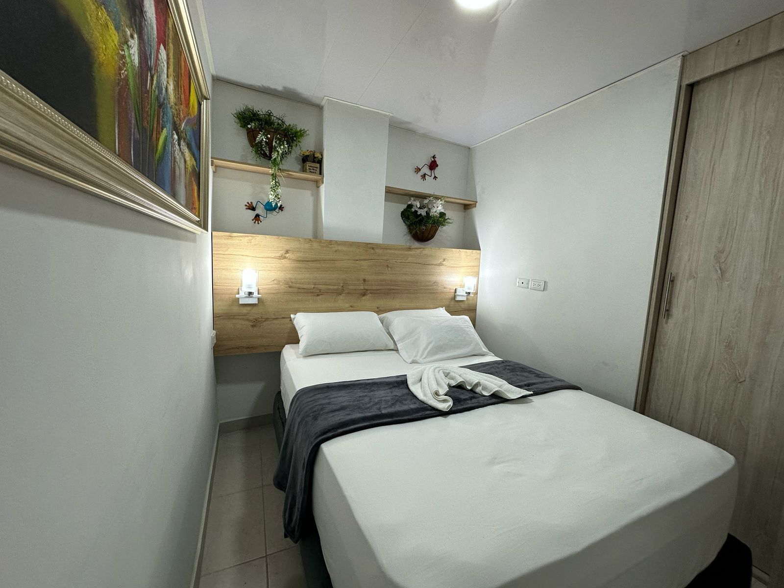 Un dormitorio con cabecera de madera, dos mesitas de noche con lámparas, cama con ropa blanca, una manta gris y estante en la pared con objetos decorativos.