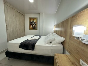 A neatly made bed with white linens and a black throw blanket, a wooden headboard, a bedside table with a lamp, and a painting on the wall. - Hospedarte Cali (Cali)