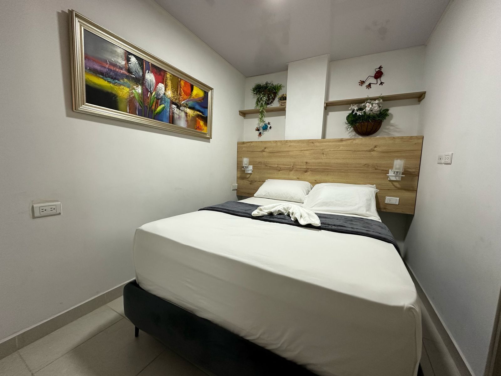 Una cama bien tendida con sábanas blancas, un cabecero de madera y un cuadro abstracto de colores encima.