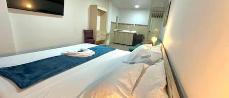 Una camera d'albergo con un letto, una coperta blu, un cuscino, un comodino, una TV a parete e un angolo cottura sullo sfondo.