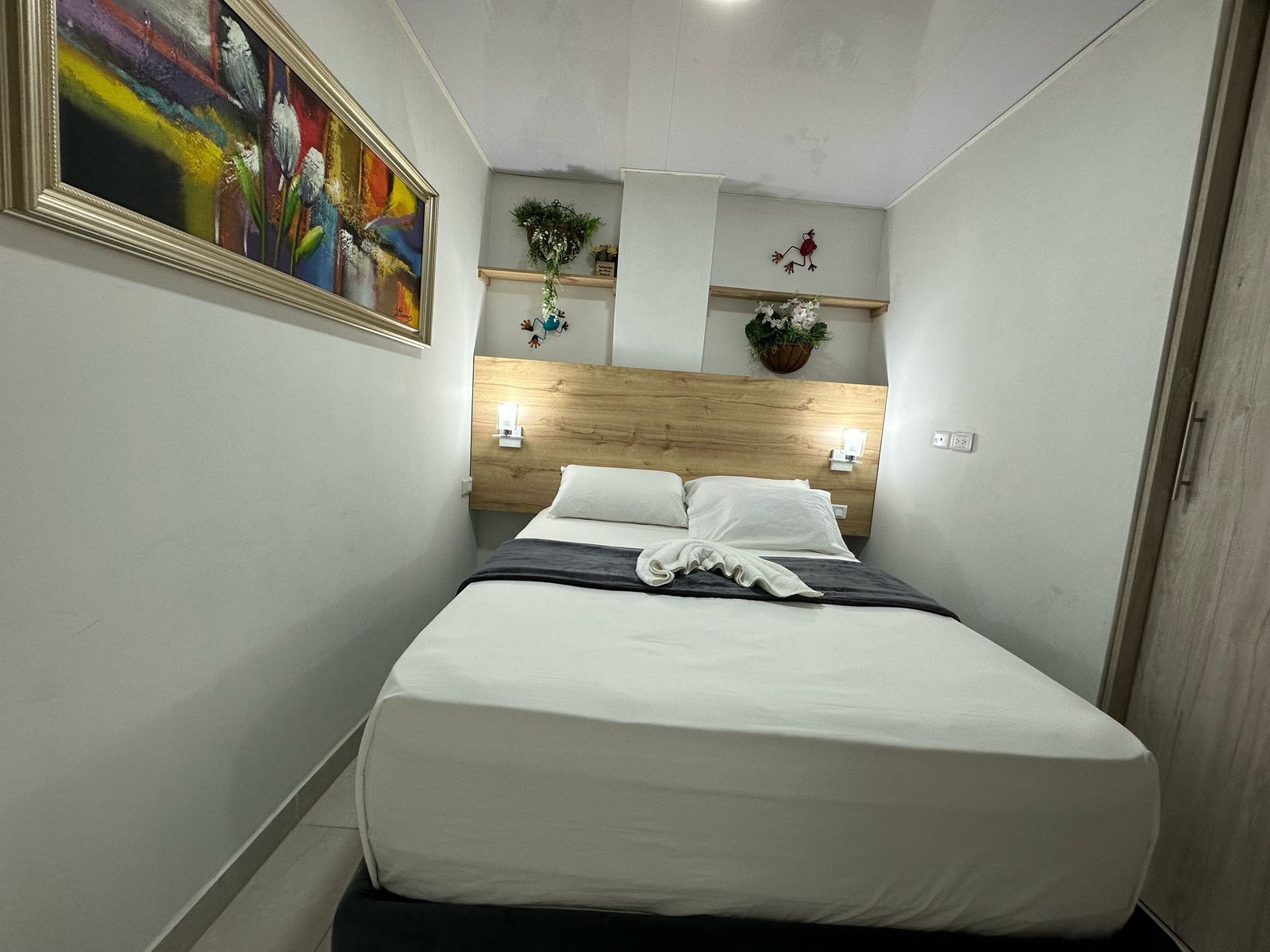 Ett litet hotellrum med en säng, en tavla på väggen och en liten växt på hyllan.