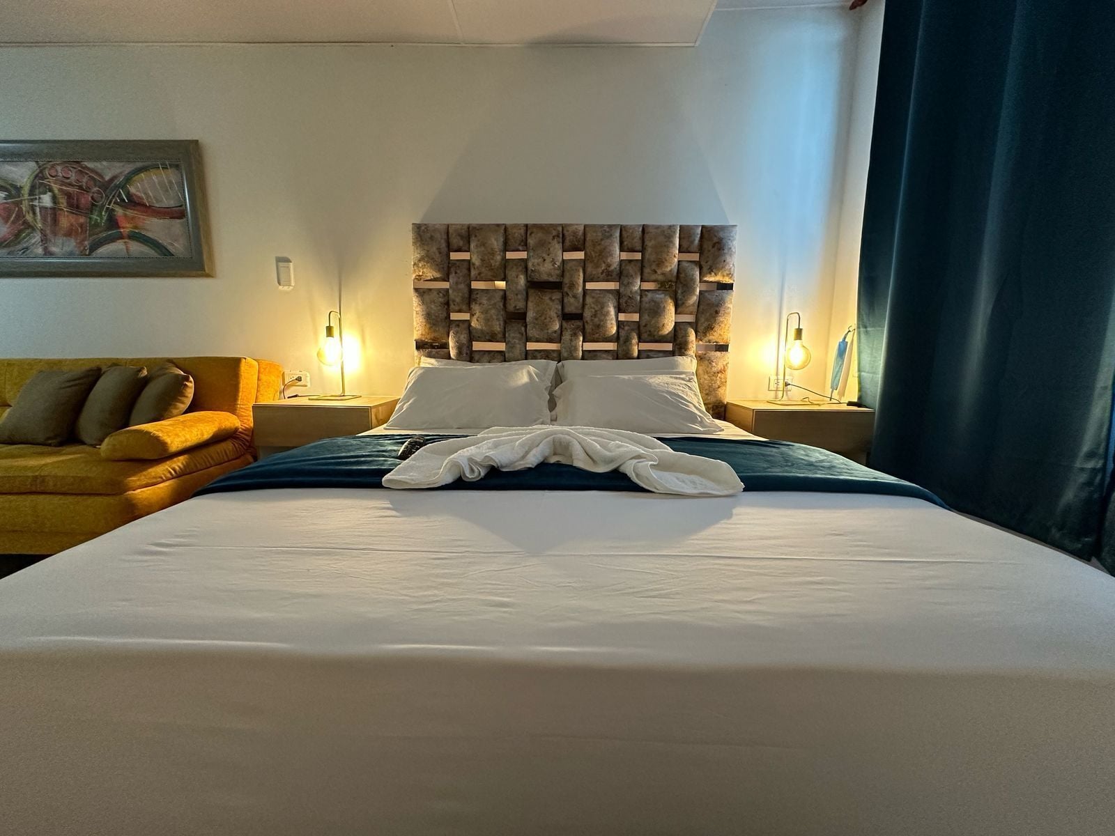 A hotel room with a large bed, a wooden headboard, a yellow sofa, and two bedside lamps.
