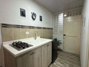 A kitchen with a gas stove, sink, and cabinets. There is a window with blinds and a door in the background. - Hospedarte Cali (Cali)