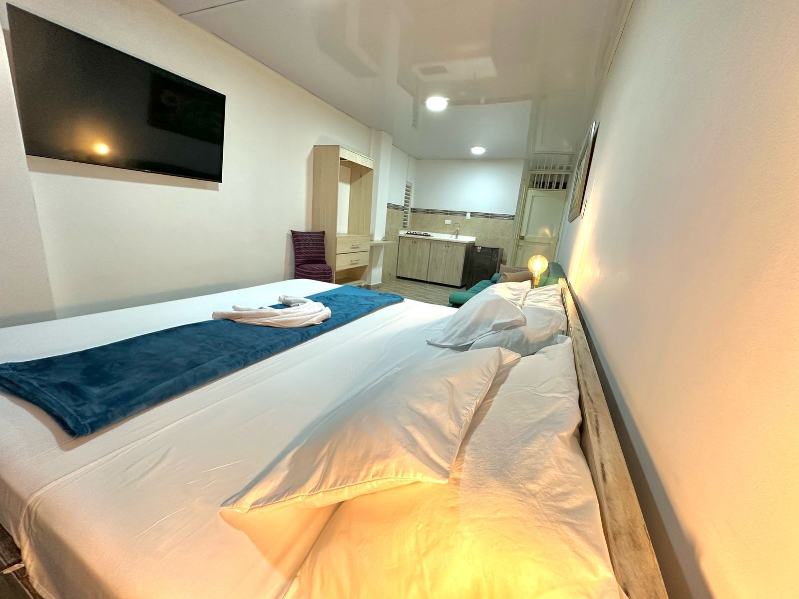 A hotel room with a bed, a blue towel, a bedside lamp, and a view of the bathroom and kitchen.