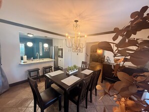 Un coin repas avec une table et des chaises noires, un lustre et une cuisine visible au fond.