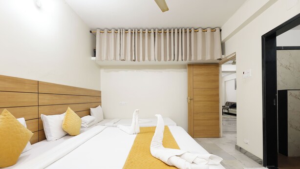 A hotel room with a large bed, wooden headboard, and a yellow accent runner. There are towels arranged on the bed, and a wooden door is visible in the background. - BALAJI HOMESTAY VARANASI (Varanasi)