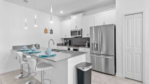 A modern kitchen with white cabinets, stainless steel appliances, a central island with bar stools, and pendant lighting. - Spacious 5BR Home With Pool Near Top Attractions (Davenport)