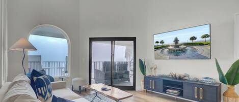 A modern living room with a sofa, a coffee table, a TV, and a balcony with outdoor furniture.