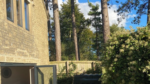 An outdoor dining area with wicker furniture and a table set for four, overlooking a stone building and a forested area. - Walnut Farm, Kingscote, Cotswolds - Unique farm stay (Kingscote)