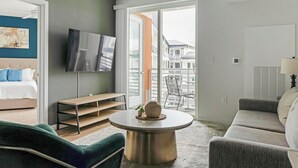 Un salon moderne avec un canapé, une table basse ronde, un téléviseur fixé au mur, un balcon offrant une vue sur les immeubles et un lit avec des coussins bleus.