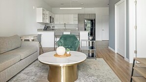 Un salon moderne avec un canapé gris, une table basse ronde surmontée d’un vase blanc et un fauteuil vert. On aperçoit à l’arrière-plan une cuisine avec des meubles blancs et des appareils en acier inoxydable.