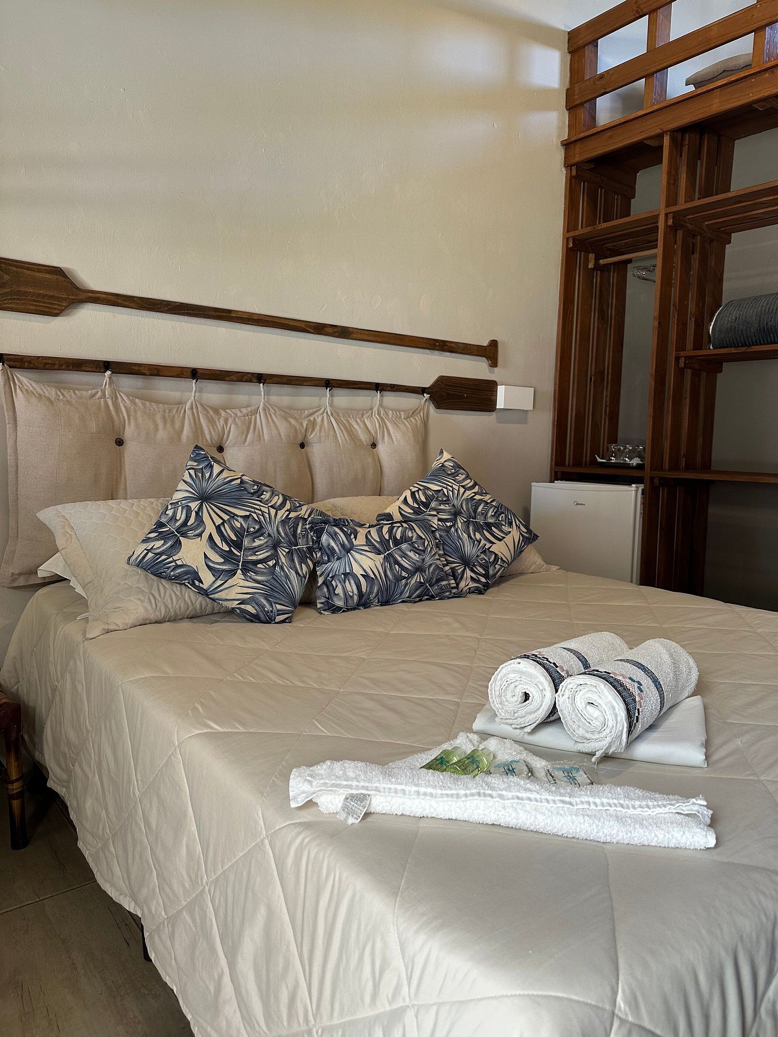 A neatly made bed with a beige quilt, two patterned pillows, and rolled towels on the bed.