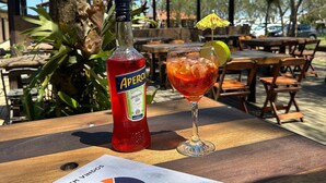 A bottle of Aperol and a glass of red drink on a wooden table outdoors with a tree and a covered area in the background. - Pousada Cardoso Surf Camping - Laguna - SC (Laguna)
