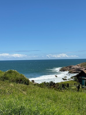 A coastal landscape with a clear view of the ocean, rocky shoreline, and a small building with a green roof. - Pousada Cardoso Surf Camping - Laguna - SC (Laguna)