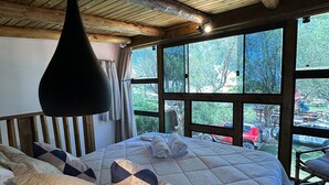 A wooden ceiling with a hanging light fixture, a large bed with a quilted duvet and pillows, and a view of the outdoors through large windows. - Pousada Cardoso Surf Camping - Laguna - SC (Laguna)